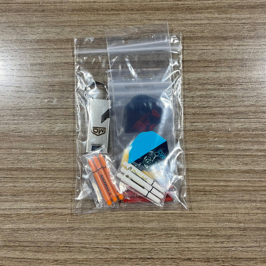 A clear plastic zip-top bag containing a small dart keychain set on a wood-grain tabletop. The set includes assorted dart Accessory Pack. Side B