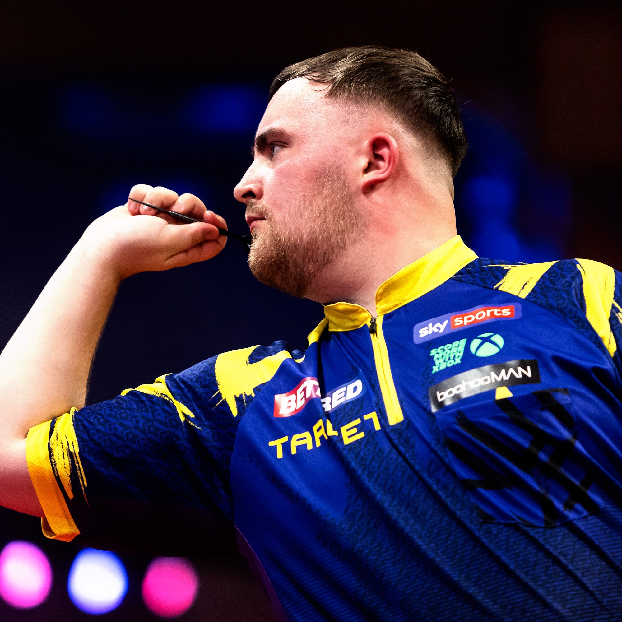 Luke Littler playing with purple and yellow sports jersey with multiple brand logos, holding a dart.