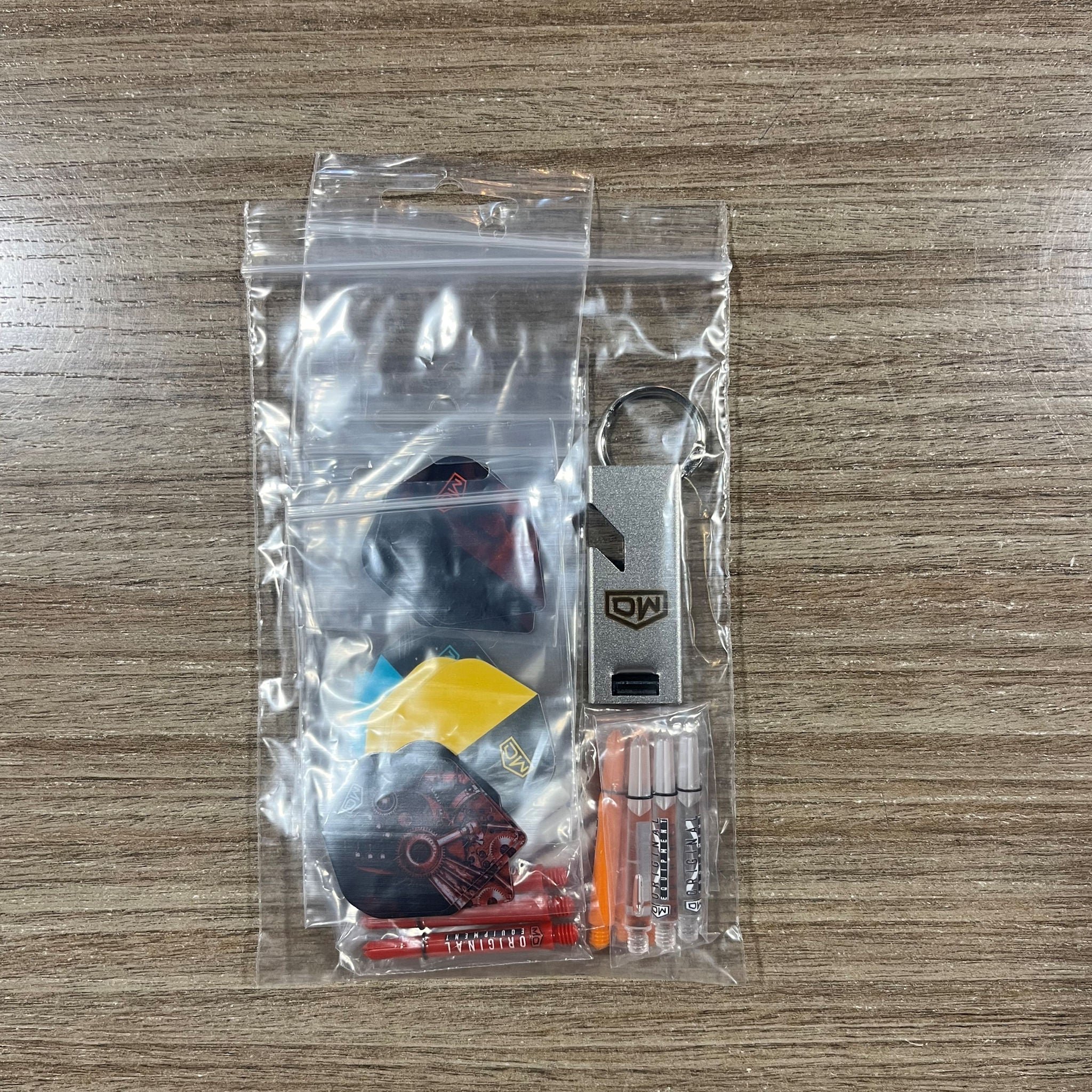 A clear plastic zip-top bag containing a small dart keychain set on a wood-grain tabletop. The set includes assorted dart Accessory Pack.  Side A