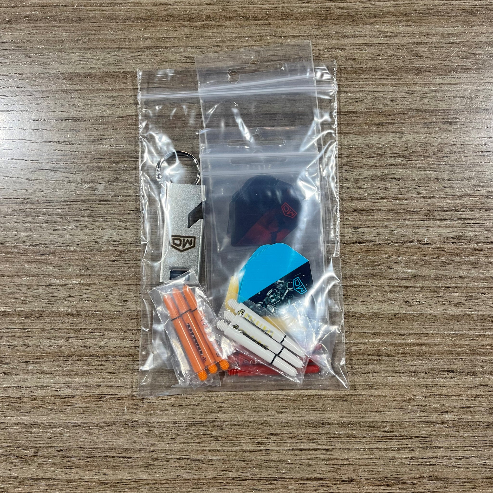 A clear plastic zip-top bag containing a small dart keychain set on a wood-grain tabletop. The set includes assorted dart Accessory Pack. Side B