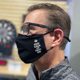 Person wearing a black face mask with 'Keep Calm and Dart On' text, in front of a dartboard.